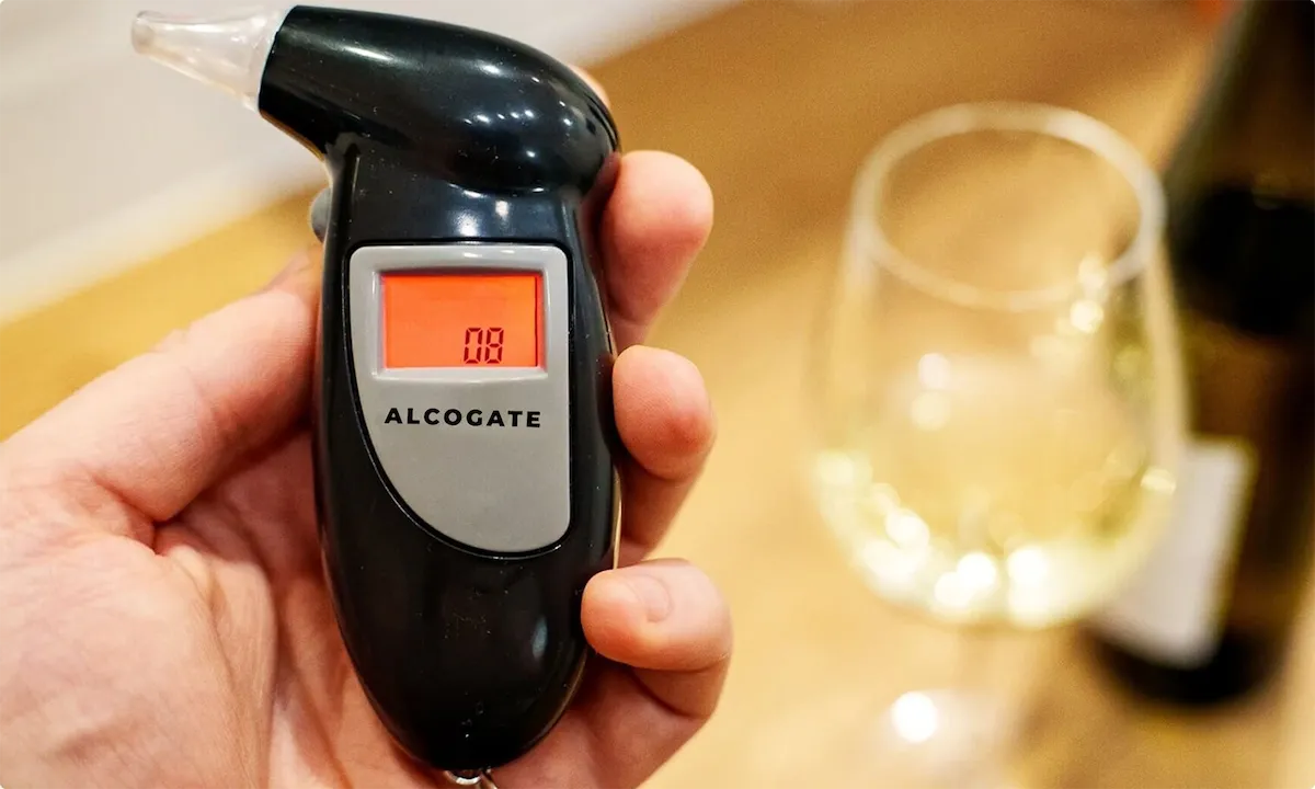 Hand holding an AlcoGate breathalyser with a glass of wine behind it